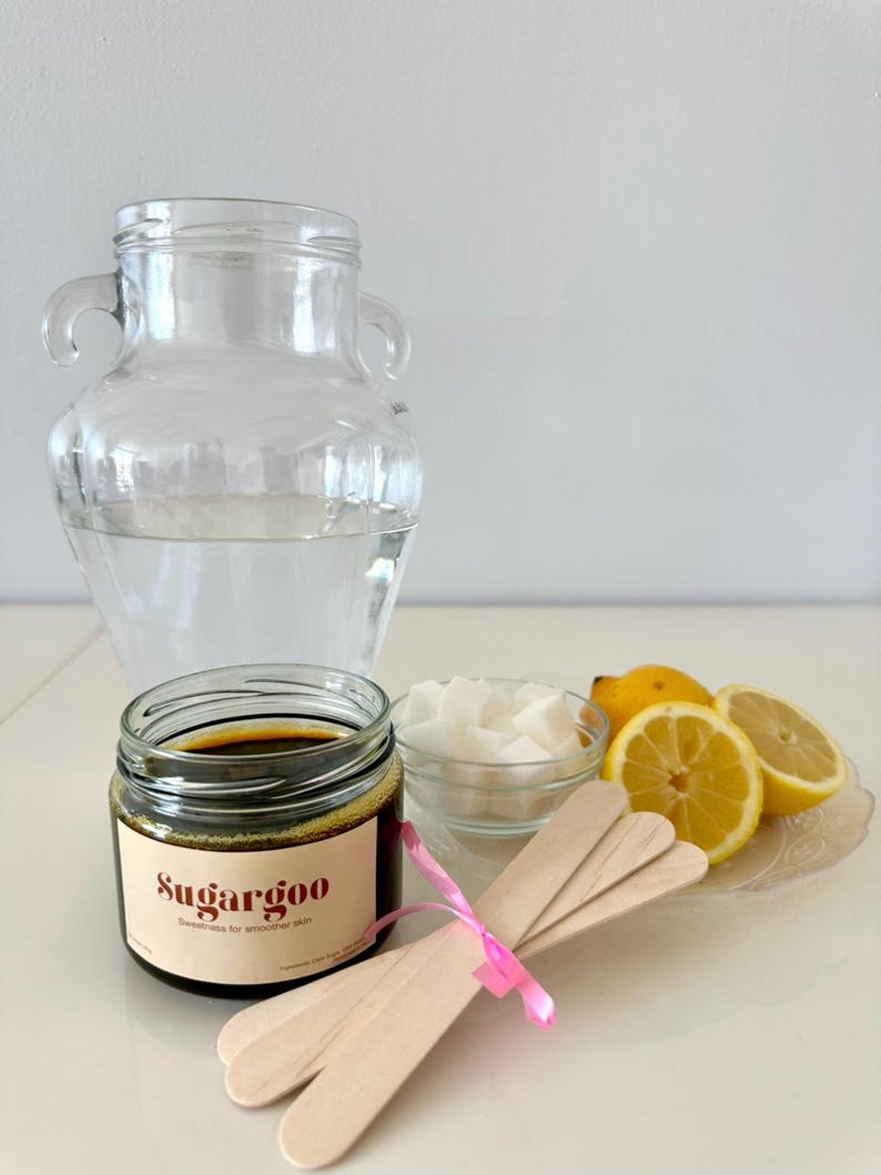 May include: A brown jar of sugar wax labeled "SugarGoo" with the tagline "Sweetness for smoother skin".  The jar is next to a bowl of white sugar cubes, a plate with three lemon wedges, and three wooden sticks tied together with a pink ribbon.