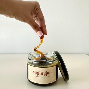 May include: A glass jar of brown sugar wax with a black lid. The jar is labeled "Sugargoo" and "Sweetness for smoother skin". A hand is holding a piece of the wax above the jar.
