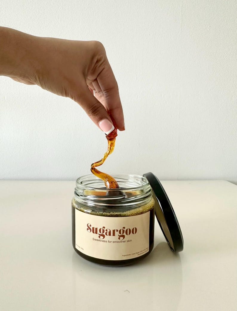 May include: A glass jar of brown sugar wax with a black lid. The jar is labeled "Sugargoo" and "Sweetness for smoother skin". A hand is holding a piece of the wax above the jar.