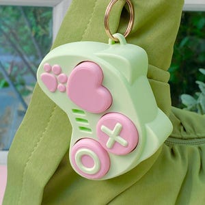 May include: A light green game controller keyring featuring pink buttons in the shape of a heart, a paw print, a plus sign, and a circle. The controller is attached to a gold-toned metal ring and clasp for easy attachment to bags or keys.