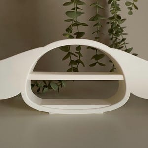 May include: A white wall shelf with a distinctive, rounded design. The shelf has two horizontal surfaces for displaying items, and a curved shape with extended sides. The shelf is made of a solid material and is set against a neutral background.