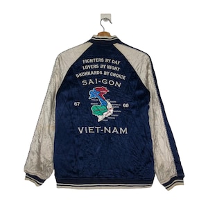 May include: A navy blue satin bomber jacket with white sleeves. The back of the jacket features an embroidered map of Vietnam with the text "FIGHTERS BY DAY LOVERS BY NIGHT DRUNKARDS BY CHOICE SAI-GON VIET-NAM".