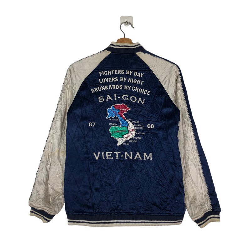 May include: A navy blue satin bomber jacket with white sleeves. The back of the jacket features an embroidered map of Vietnam with the text "FIGHTERS BY DAY LOVERS BY NIGHT DRUNKARDS BY CHOICE SAI-GON VIET-NAM".