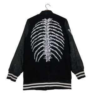 May include: Black varsity jacket with white rib cage graphic on the back. The jacket has black sleeves, white and black striped cuffs and hem. The jacket is hanging on a wooden hanger.