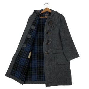 May include: A gray wool duffel coat with a hood and toggle closures. The coat has a blue and black plaid lining and two front pockets. The coat is hanging on a wooden hanger.
