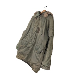 Op de afbeelding: Olijfgroene parka met een met bont gevoerde capuchon, hangend aan een houten hanger. De jas heeft meerdere zakken, een ritssluiting en een patch op de bovenarm. De jas is in vintage stijl.