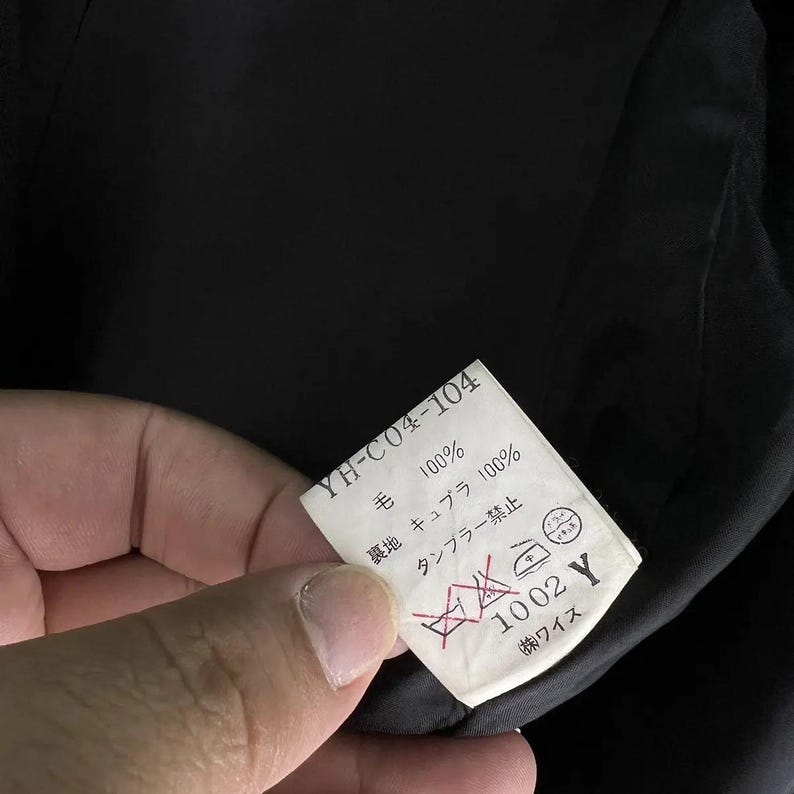 May include: A white tag with black text and care symbols is held up against a black garment. The tag reads "YH-C04-104", "100%", "キュプラ 100%", and "1002 Y".