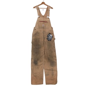 Könnte beinhalten: Eine beige Arbeitshose mit einem Skelett-Hand-Patch auf der rechten Tasche. Die Hose ist abgenutzt und hat Schmutzflecken.