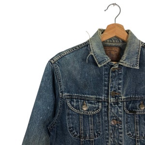 Puede incluir: Una chaqueta vaquera azul con una etiqueta de cuero marr&oacute;n que dice "Edwin Co. Ltd.". La chaqueta tiene cierre de botones y dos bolsillos delanteros.