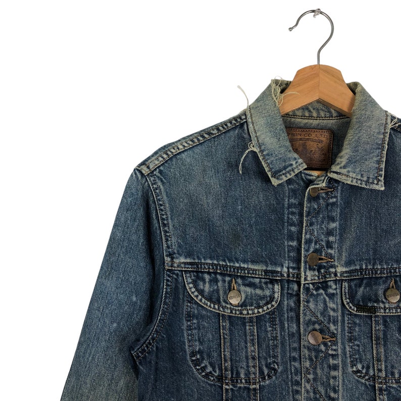 Puede incluir: Una chaqueta vaquera azul con una etiqueta de cuero marr&oacute;n que dice "Edwin Co. Ltd.". La chaqueta tiene cierre de botones y dos bolsillos delanteros.