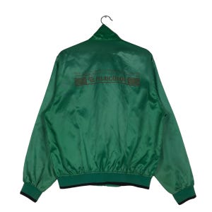 Puede incluir: Una chaqueta bomber de satén verde con cremallera negra y puños y cintura de canalé negros. La parte posterior de la chaqueta tiene el texto "YOU'LL GET SUPERIOR QUALITY AND PERFORMANCE FROM OUR TECHNOLOGY. WE'RE ALWAYS TRYING TO MAKE IT BETTER." y el logotipo "FUJICOLOR" en marrón.