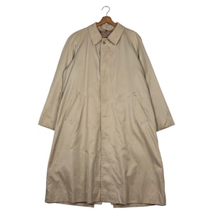 Könnte beinhalten: Ein beigefarbener Trenchcoat mit klassischem Design, mit Kragen, Knopfverschluss und zwei Vordertaschen. Der Mantel hängt an einem Holzbügel vor weißem Hintergrund. Der Mantel ist aus einem glatten, strapazierfähigen Stoff gefertigt.