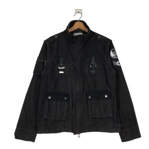 Könnte beinhalten: Schwarze Jacke mit mehreren Taschen, Schnallen und Stehkragen. Die Jacke hat einen Aufnäher mit Totenkopf und Flügeln am Oberarm. Die Jacke hängt an einem Holzbügel.