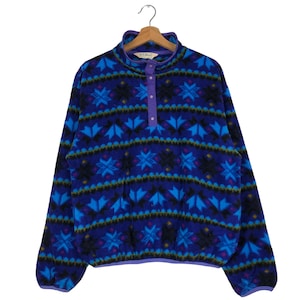 Op de afbeelding: Een blauwe fleece trui met een geometrisch patroon van blauwe, zwarte en paarse vormen. De trui met lange mouwen heeft een opstaande kraag met drie paarse knopen en een paarse bies. Het L.L.Bean-logo is zichtbaar op de kraag.