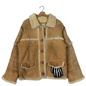 May include: A tan shearling jacket with a cream-colored shearling collar and trim. The jacket has a button-up front, two front pockets, and a black and white skeleton hand patch on the pocket. The jacket is hanging on a wooden hanger.
