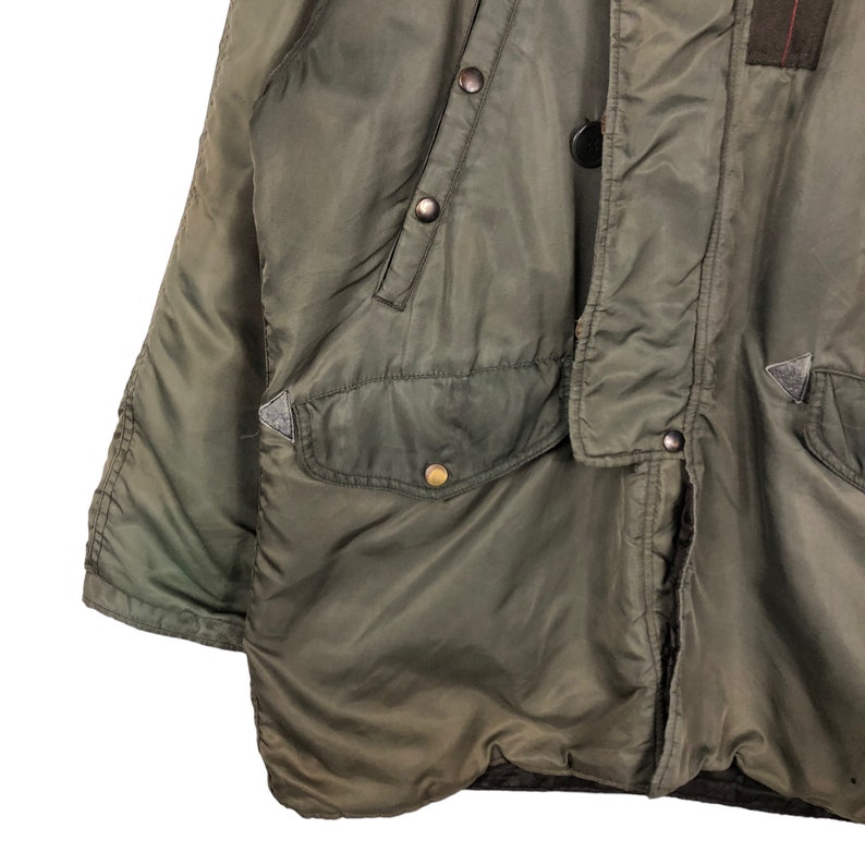 Op de afbeelding: Close-up van een olijfgroene winterjas. De jas heeft een gewatteerd ontwerp, meerdere zakken en knoopsluitingen. De jas heeft een militair ge&iuml;nspireerde esthetiek, met een duurzaam, weerbestendig materiaal.