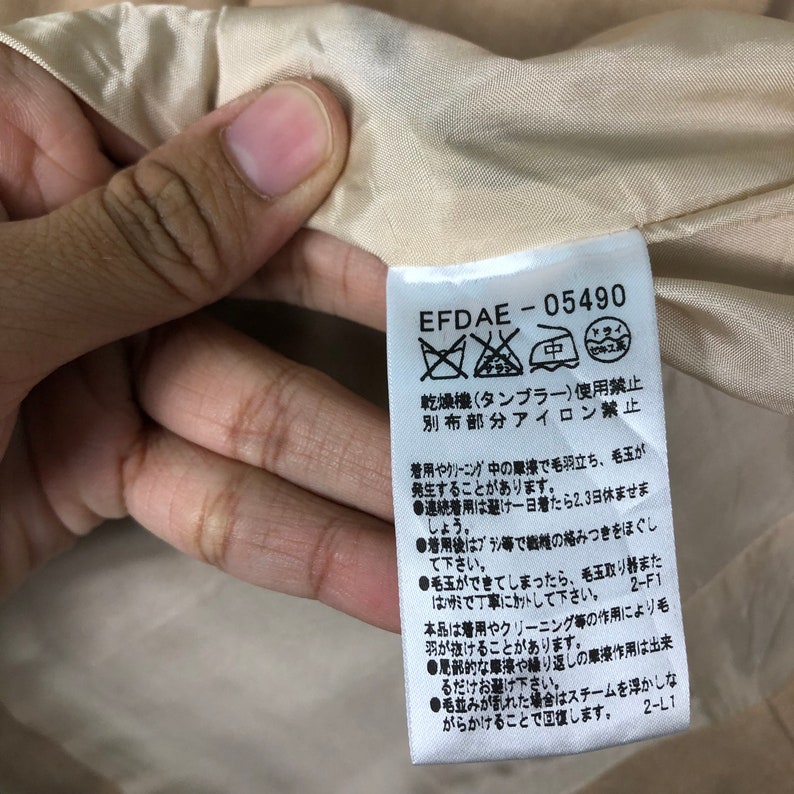 Op de afbeelding: Close-up van een label met de tekst "EFDAE-05490" en Japanse karakters. Het label is wit met zwarte tekst en is bevestigd aan een beige stof. Het label geeft onderhoudsinstructies.