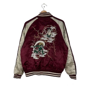 May include: A maroon and cream satin souvenir jacket featuring long sleeves with cream panels. The back displays an embroidered design of two green and white mythical figures surrounded by white clouds and yellow lightning bolt patterns.