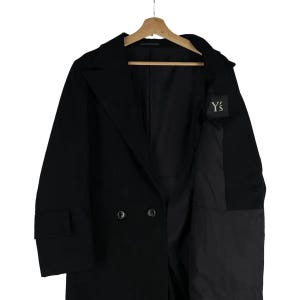 May include: A black double-breasted coat with a notched lapel, hanging on a wooden hanger. The coat has two rows of buttons and a small label with the text "Y's". The coat is made of a dark fabric.