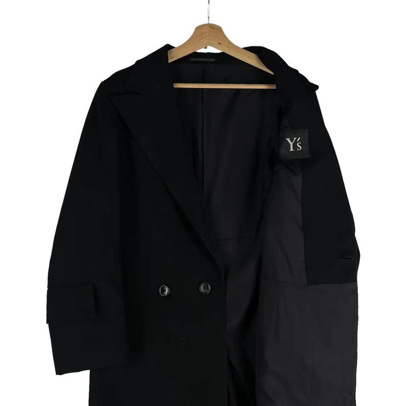 May include: A black double-breasted coat with a notched lapel, hanging on a wooden hanger. The coat has two rows of buttons and a small label with the text "Y's". The coat is made of a dark fabric.