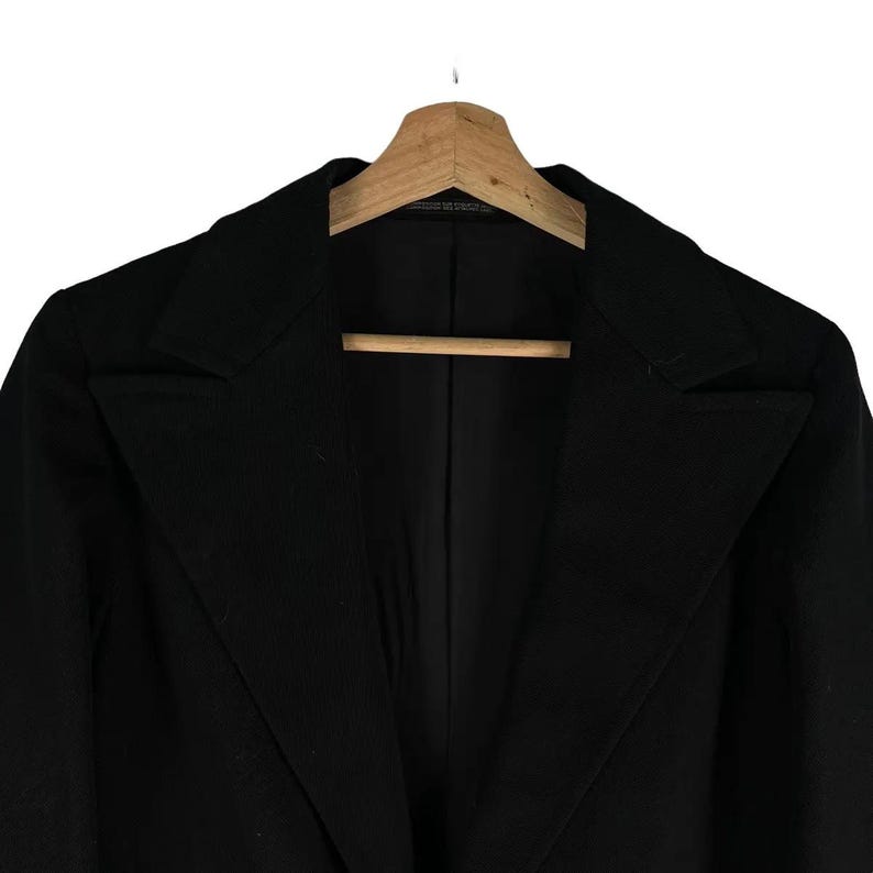 May include: A black blazer with a notched lapel, hanging on a wooden hanger. The jacket has a classic, tailored design with a single-breasted closure. The fabric appears to be a smooth, solid material.