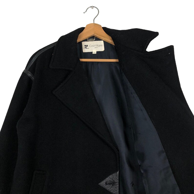 May include: A black wool coat with leather accents on the shoulders and a notched collar. The coat is hanging on a wooden hanger. The interior lining is a dark blue color. The coat has a label that reads "Courr&egrave;ges".