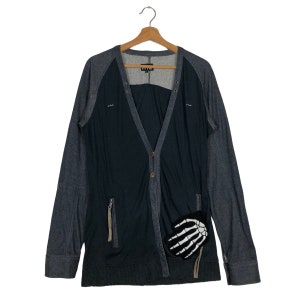 May include: A dark gray and black cardigan with long sleeves, a V-neck, and a button-up front. The cardigan features a skeleton hand patch on the pocket and a zipper pocket. The garment is hanging on a wooden hanger.