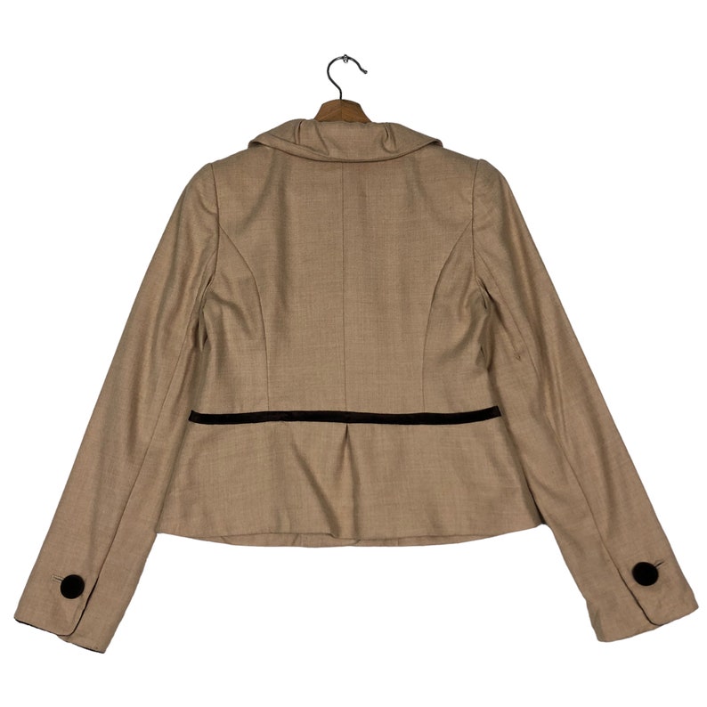 Op de afbeelding: Beige blazer met zwarte bies en zwarte knopen op de mouwen, hangend aan een houten kleerhanger. De achterkant van de jas is zichtbaar tegen een witte achtergrond. Klassiek ontwerp.
