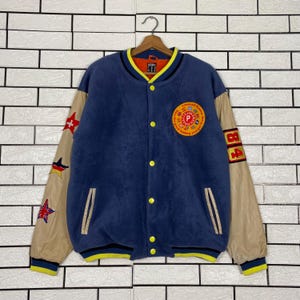 May include: A blue fleece varsity jacket with beige sleeves, yellow snap buttons, and a yellow-trimmed collar. The left chest features a circular patch with the text "Person's" and "The good feeling you get from". The right sleeve has star patches, and the left sleeve has the number "84" in red and yellow.