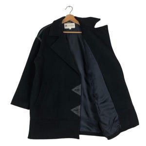 May include: A black wool coat with a notched collar and a dark blue interior lining. The coat features leather accents on the shoulders and decorative leather triangles near the hem. The coat is hanging on a wooden hanger.
