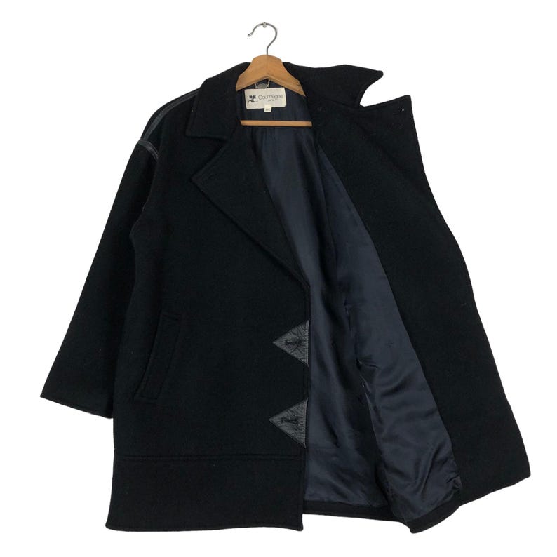 May include: A black wool coat with a notched collar and a dark blue interior lining. The coat features leather accents on the shoulders and decorative leather triangles near the hem. The coat is hanging on a wooden hanger.