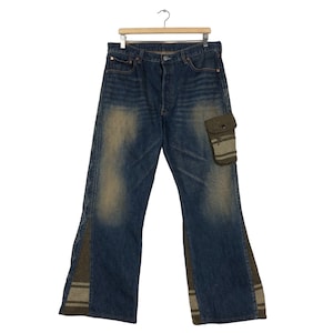 May include: Blue denim jeans with a faded wash and unique fabric accents. Features a small pocket on the right leg and patterned fabric inserts on the sides. The jeans are hanging on a wooden hanger.
