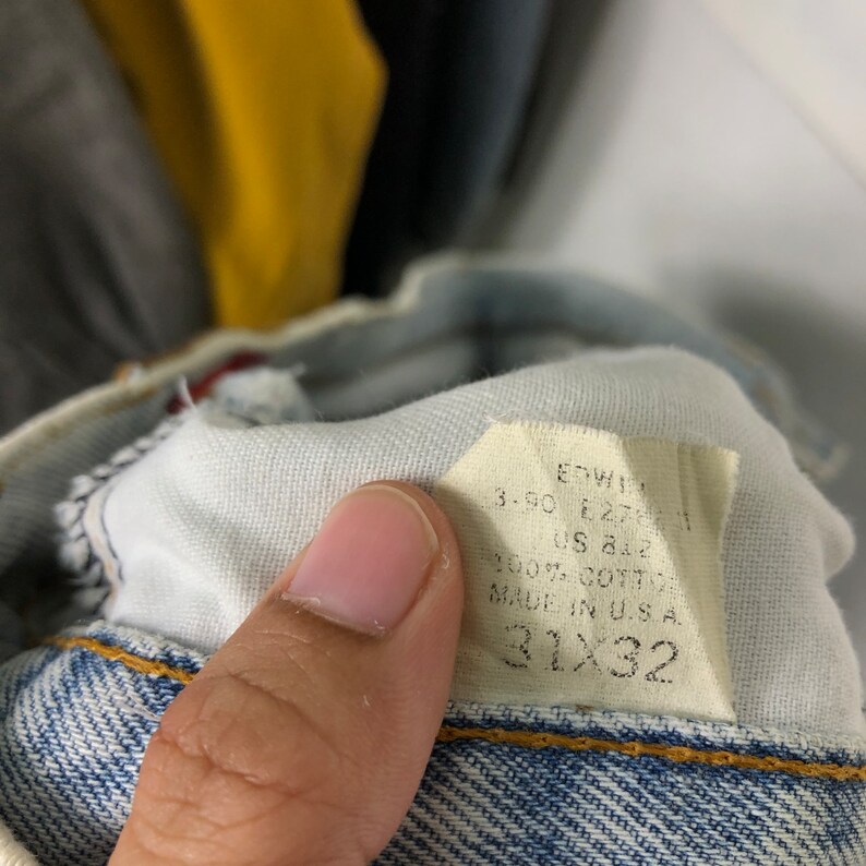 Może przedstawiać: Biała metka z czarnym tekstem, na kt&oacute;rej widnieje napis "Edwin 3.50 E.27-1-1 US A12 100% Bawełna Wyprodukowano w USA 31x32", przymocowana do pary jasnoniebieskich dżins&oacute;w.