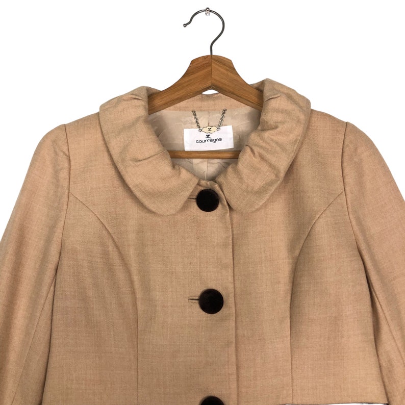 Op de afbeelding: Een beige jasje met een ronde kraag en drie grote, donkerbruine knopen. De jas hangt aan een houten kleerhanger. Het label luidt "courreges".