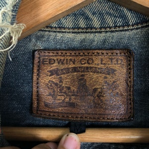Puede incluir: Un parche de cuero marr&oacute;n con el texto "EDWIN CO. LTD" y un logotipo con una corona y el texto "WEST INDIES" en un color azul descolorido.