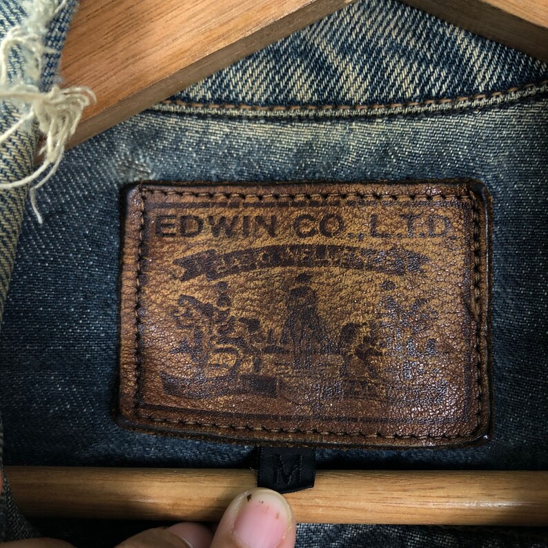 Puede incluir: Un parche de cuero marr&oacute;n con el texto "EDWIN CO. LTD" y un logotipo con una corona y el texto "WEST INDIES" en un color azul descolorido.