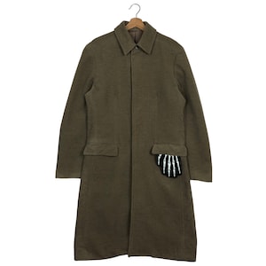 May include: Olive green long coat with a collared neckline and two front pockets. A black and white skeleton hand patch is on the right pocket. The coat is hanging on a wooden hanger.