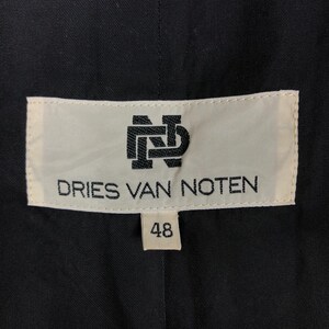 Może przedstawiać: Biała etykieta z czarnym tekstem, kt&oacute;ry brzmi "DRIES VAN NOTEN" i logo z przeplatanymi literami "DN". Etykieta ma r&oacute;wnież mniejszą białą etykietę z numerem "48" na niej.