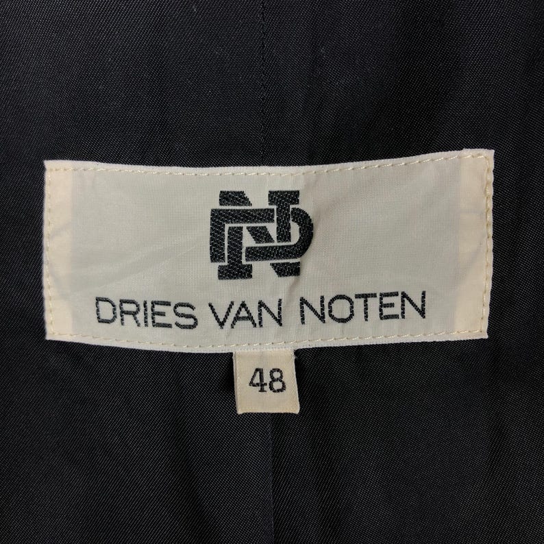 Może przedstawiać: Biała etykieta z czarnym tekstem, kt&oacute;ry brzmi "DRIES VAN NOTEN" i logo z przeplatanymi literami "DN". Etykieta ma r&oacute;wnież mniejszą białą etykietę z numerem "48" na niej.