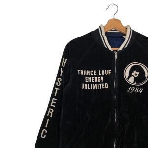 May include: Black velvet bomber jacket with white embroidered lettering on the sleeves and back. The lettering reads "HYSTERIC" on the sleeves and "TRANCE LOVE ENERGY UNLIMITED 1984" on the back. The jacket has a zipper closure and a ribbed collar.