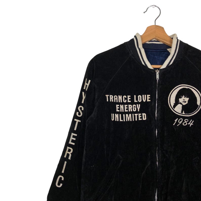 May include: Black velvet bomber jacket with white embroidered lettering on the sleeves and back. The lettering reads "HYSTERIC" on the sleeves and "TRANCE LOVE ENERGY UNLIMITED 1984" on the back. The jacket has a zipper closure and a ribbed collar.