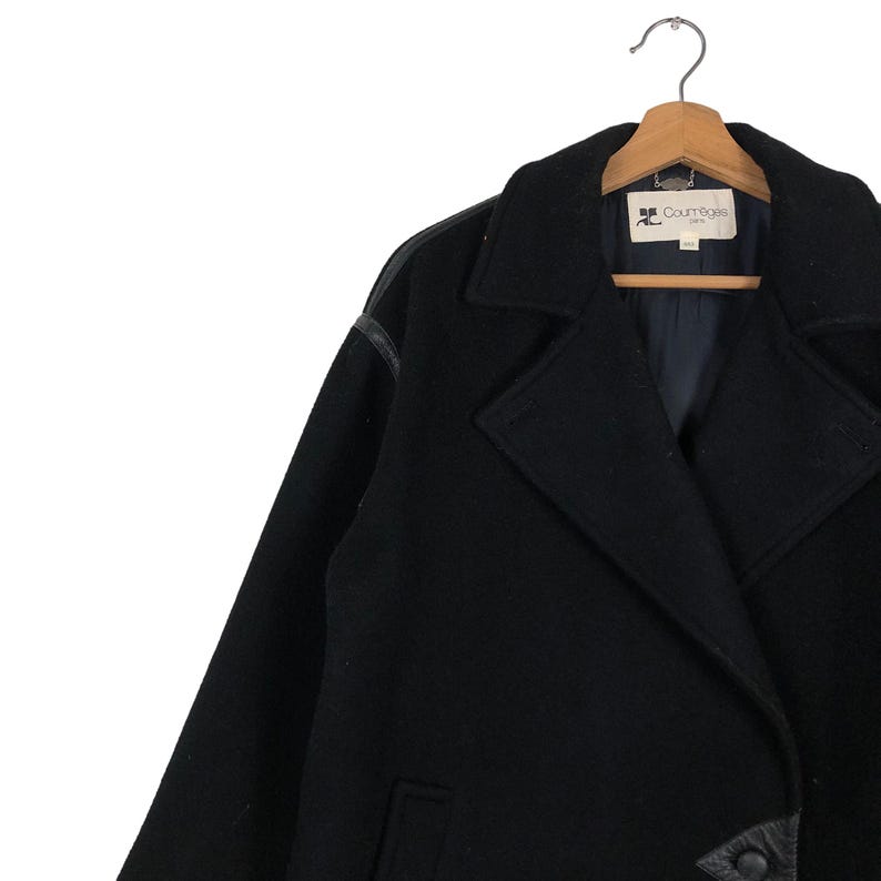 May include: A black wool coat with a notched collar and leather accents on the shoulders. The coat has a double-breasted design and a small pocket. The label reads "Courr&egrave;ges PARIS". The coat is hanging on a wooden hanger.