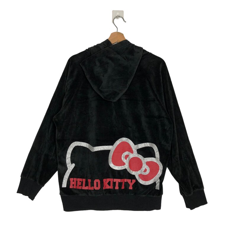 May include: Black velour hoodie with a large Hello Kitty graphic on the back. The graphic is silver with red and pink accents. The hoodie has a hood and a zipper.