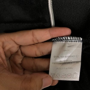 May include: A white tag with black text attached to a black garment. The tag reads "QIN-14 &copy;'76, '14 SANRIO (株)サザック 大阪市中央区安土町3-4-16 ポリエステル100%"