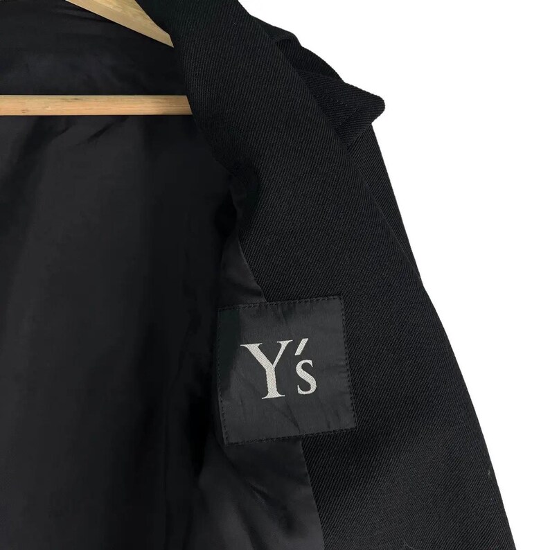 May include: Black coat with a visible interior lining. The coat features a label with the text "Y's" in a stylized font. The coat is hanging on a wooden hanger. The fabric appears to be a woven material.