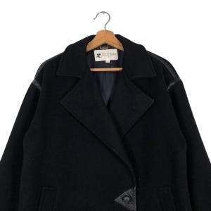 May include: Black wool coat with leather accents on the shoulders and a double-breasted closure. The coat features a notched collar and is hanging on a wooden hanger. The Courr&egrave;ges logo is visible on the label.