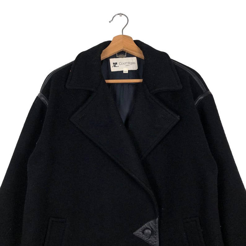 May include: Black wool coat with leather accents on the shoulders and a double-breasted closure. The coat features a notched collar and is hanging on a wooden hanger. The Courr&egrave;ges logo is visible on the label.