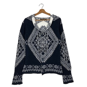 Könnte beinhalten: Eine marineblaue Strickjacke mit weißem geometrischen Muster. Der Pullover hat einen Schalkragen und einen Knopfverschluss. Das Design zeigt Rauten- und Zickzackmuster. Die Strickjacke hängt an einem Holzbügel.