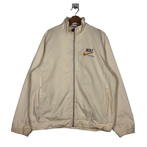 May include: A cream-colored zip-up jacket with a black zipper and a Nike logo on the left chest. The logo is orange and black with the text "NIKE SPORTSWEAR" underneath.