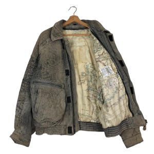 Könnte beinhalten: Eine verwitterte graue Lederjacke mit einem kartenbedruckten Futter. Die Jacke hat eine strukturierte Außenseite, mehrere Taschen und einen gerippten Saum. Sie hängt an einem Holzbügel vor weißem Hintergrund.